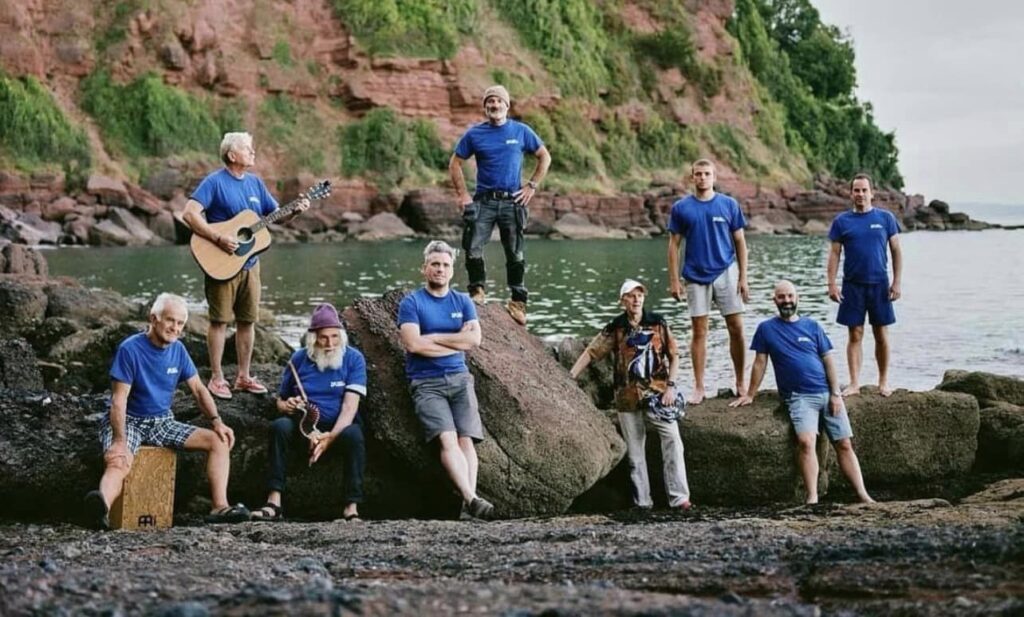 The Out of Tunas shanty band, wearing blue shirts and sat on rocks on a beach.