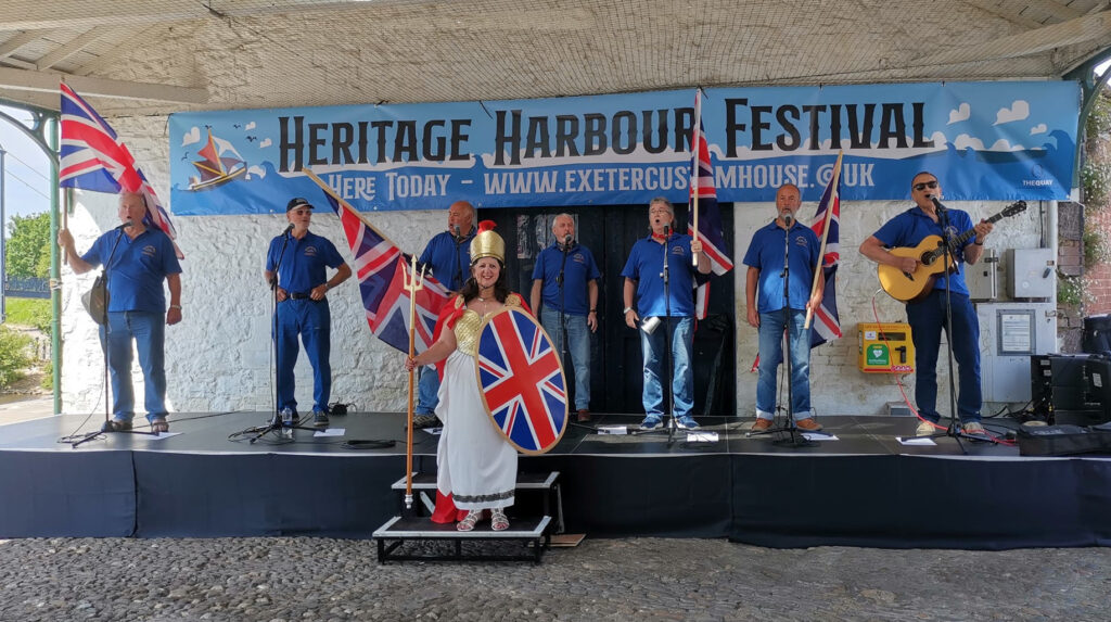 Mariner's Away shanty band singing under the Transit Shed wearing blue shirts and holding union jack flags. In the middle is a women dressed as Britannia, with a union jack shield.