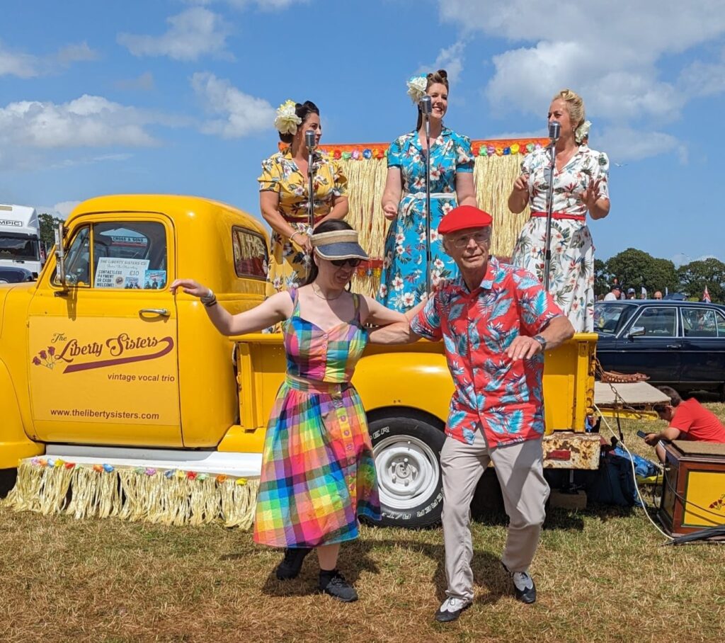 South West Lindy Hoppers dancing in front of a yellow pick-up truck where the three girls from the Liberty Sisters are all performing. They are all wearing really bright summery clothes.