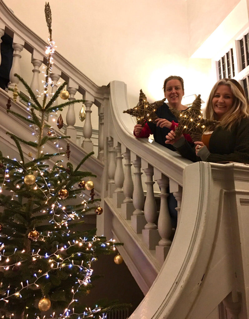 Two women holiday willow stars they have made on the staircase inside the Custom House. There is a parge Christmas tree besides them.