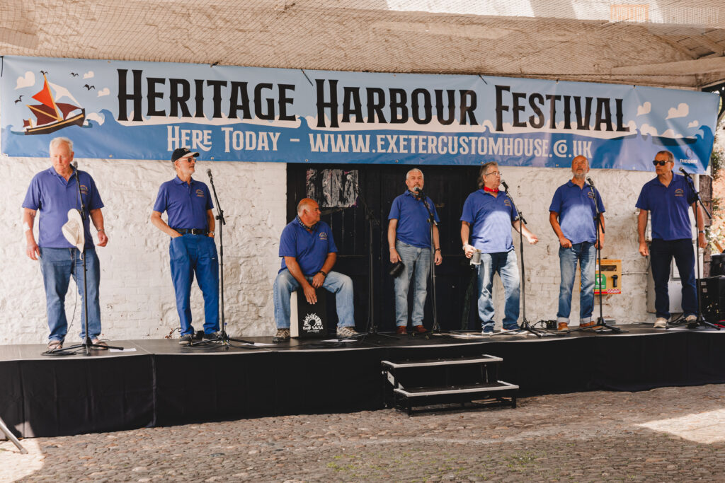Photo of five members of Mariner's Away shanty band performing on a stage under the Transit Shed. They are all wearing blue smocks and shirts and are standing in front of a banner for the festival.