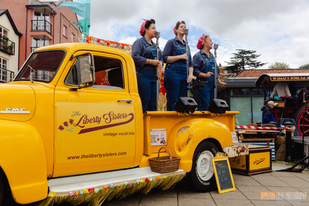 The three members of the Liberty Sisters performing on the back of a yellow American-style pickup truck. They are dressed in 1940's era blue dungarees.