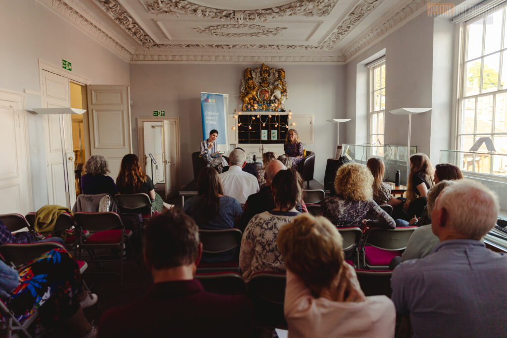 Audience sitting in the Long Room on chairs listening to a talk by Literature Works, with two women sat on stage doing an interview.